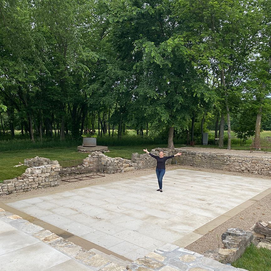 Woman with arms outstretched in ancient ruins, stone walls, trees, green field.