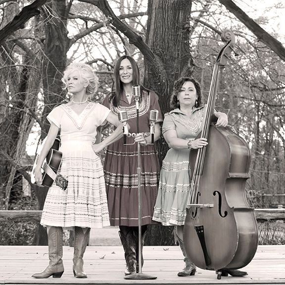 Three women in vintage outfits posing outdoors with a guitar, microphone, and upright bass.