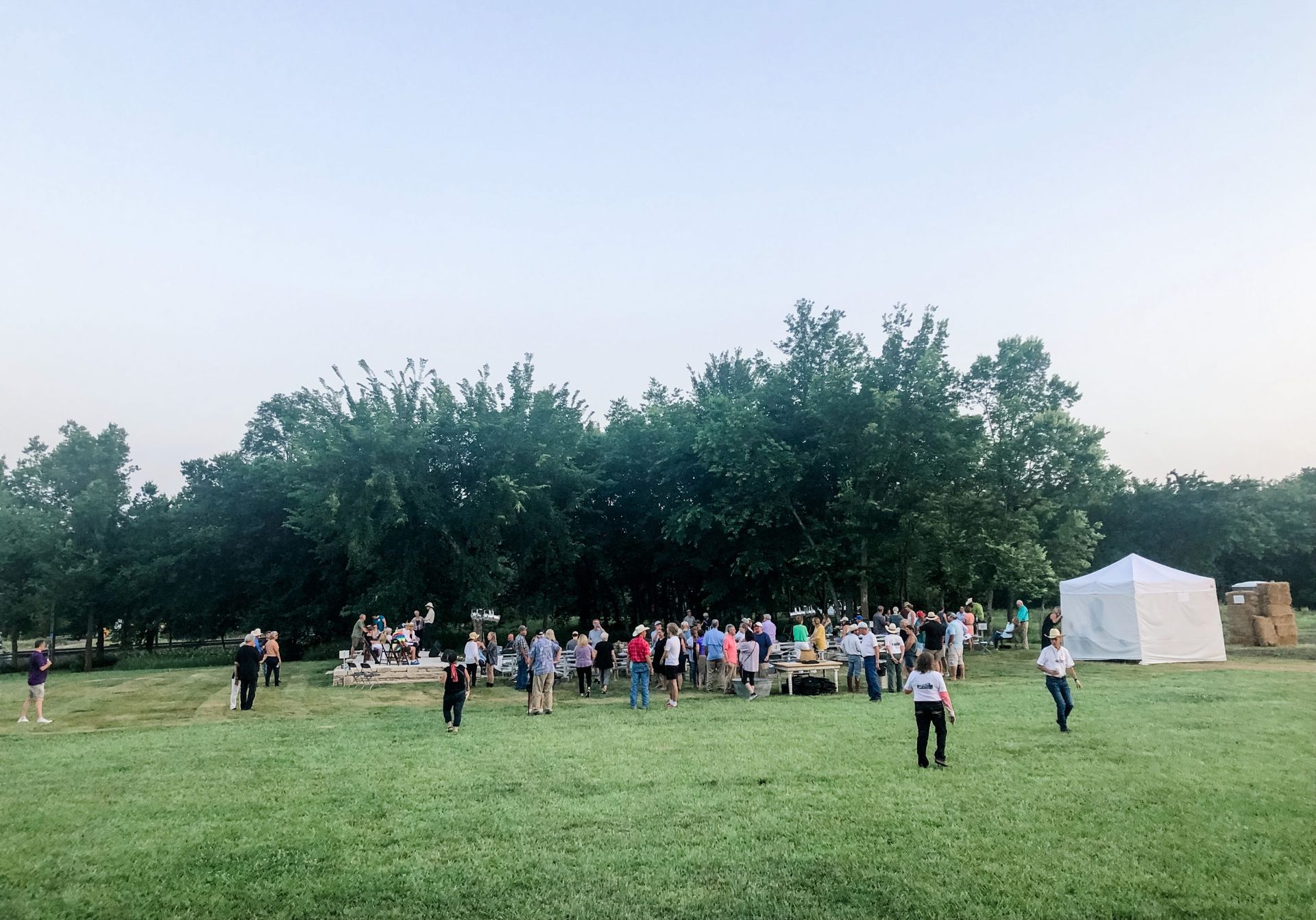 People gather outdoors on a grassy field near a treeline and a white tent.