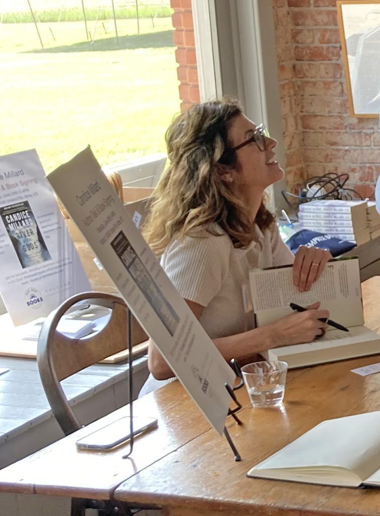 Woman at a table signing books. She wears glasses and smiles, light-filled room, posters and books present.
