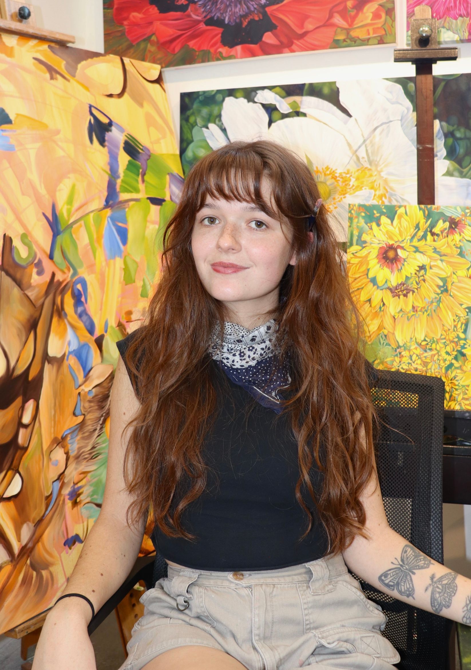 Woman with long brown hair in artist's studio, surrounded by paintings of flowers.