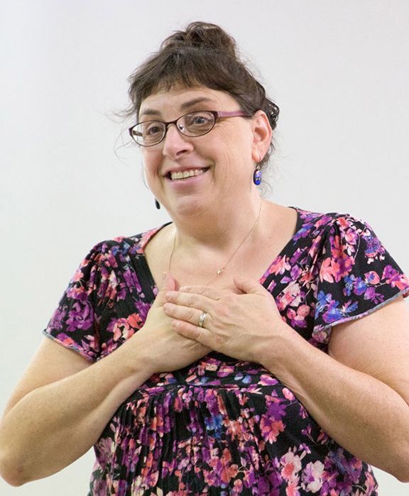 A woman wearing glasses and a floral shirt is smiling with her hands on her chest