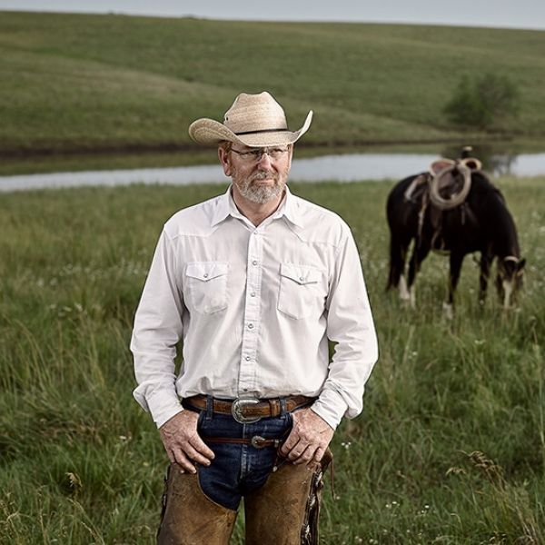A man in a cowboy hat is standing in a field with two horses in the background