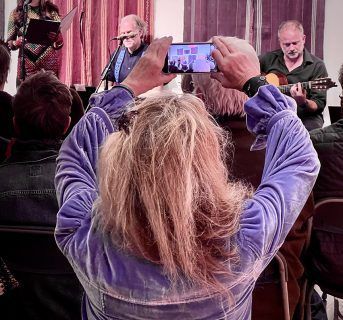Woman in blue velvet jacket taking photo of a band performing onstage.