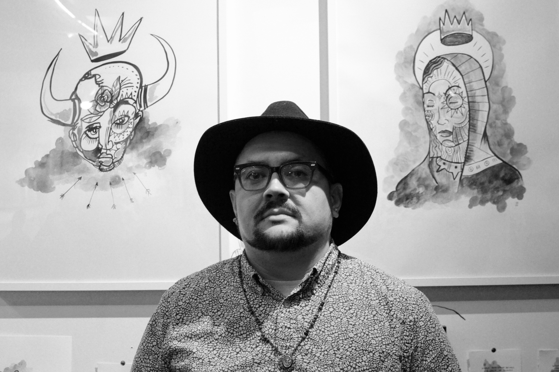 A man wearing a hat and glasses is standing in front of a wall with drawings on it.