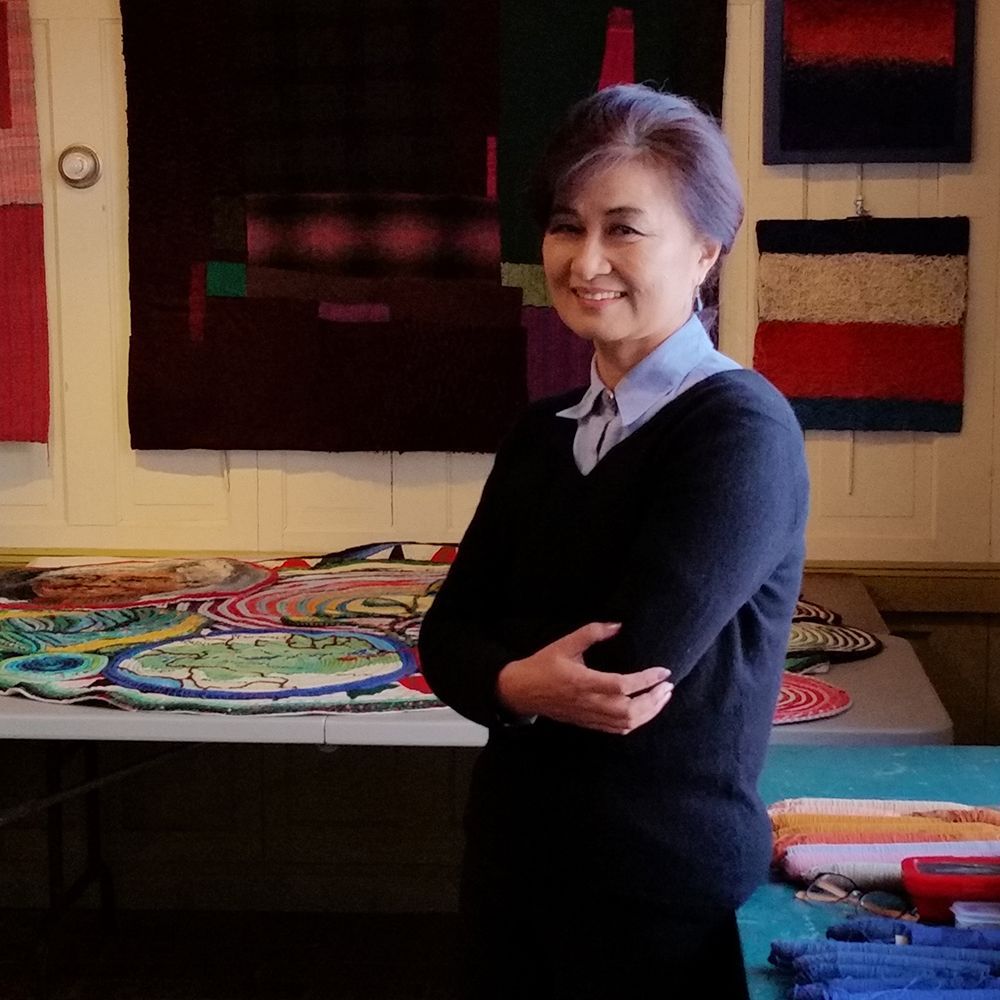 A woman in a black sweater is standing in front of a table with quilts on it