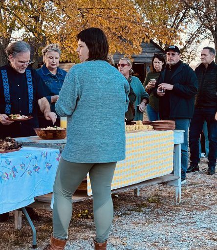 People at an outdoor gathering, serving food from tables. Several in casual wear, some eating.