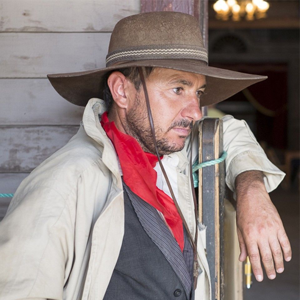 A man wearing a hat and a red scarf leans against a wall