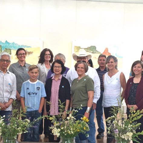 Group of people standing in front of colorful artwork, some holding flowers, smiling.