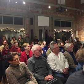 Audience in a dimly lit hall watches a presentation. People sit, listen, and look toward a screen.