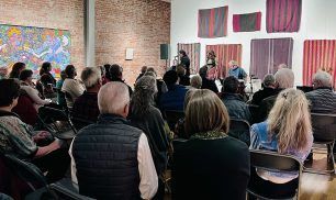 Audience at an art gallery event, listening to two people on a stage. Art on walls.