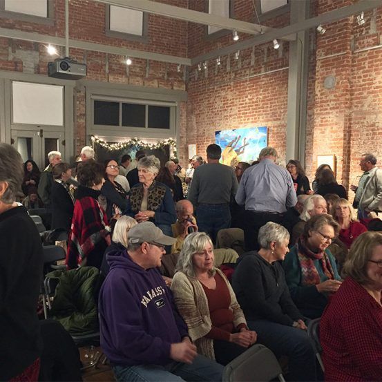 People gathered in a gallery with brick walls, some seated, viewing art.