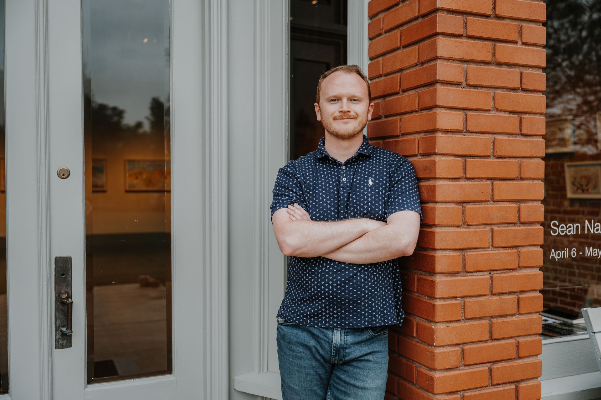 Man with arms crossed, leans against brick column by a white door. He wears a blue shirt and jeans.