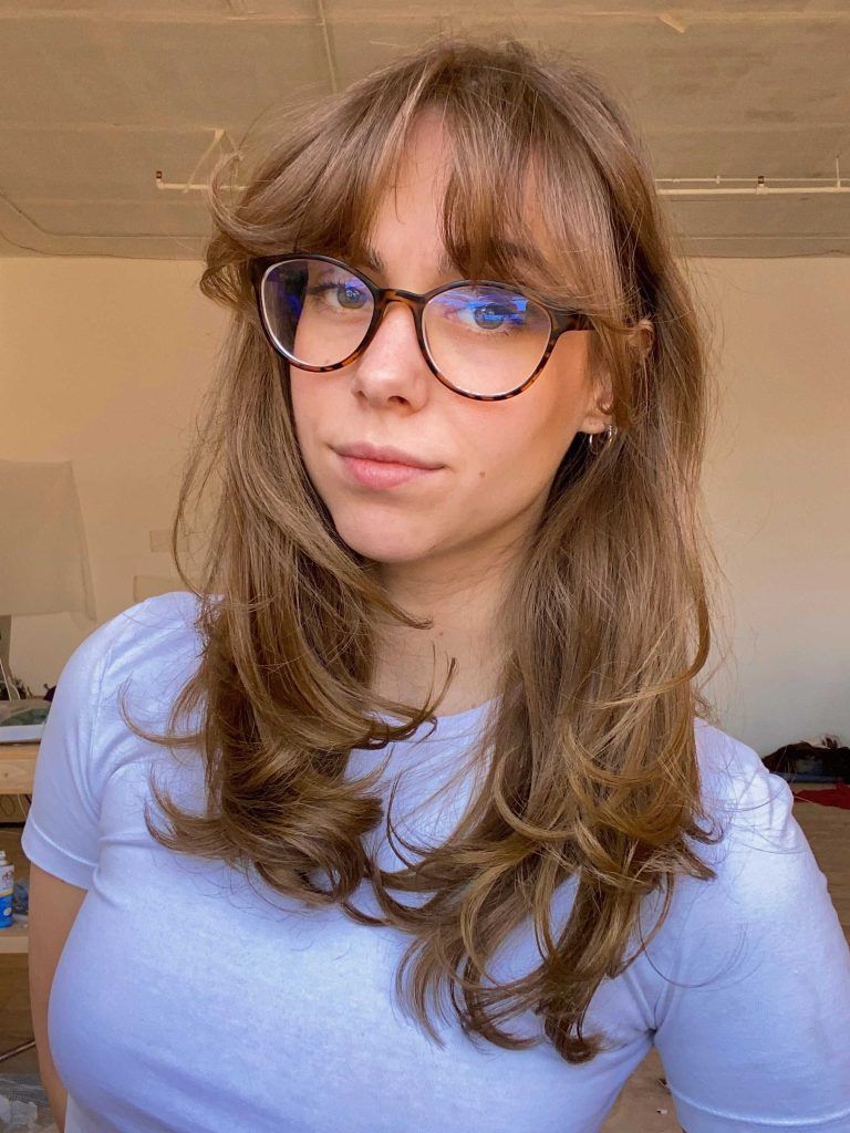 Woman with glasses, light brown hair, and white t-shirt looking at the camera. Indoor setting, natural light.