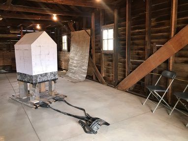 Indoor scene: white house-shaped sculpture on a platform with black cables in a rustic wooden building with two chairs.