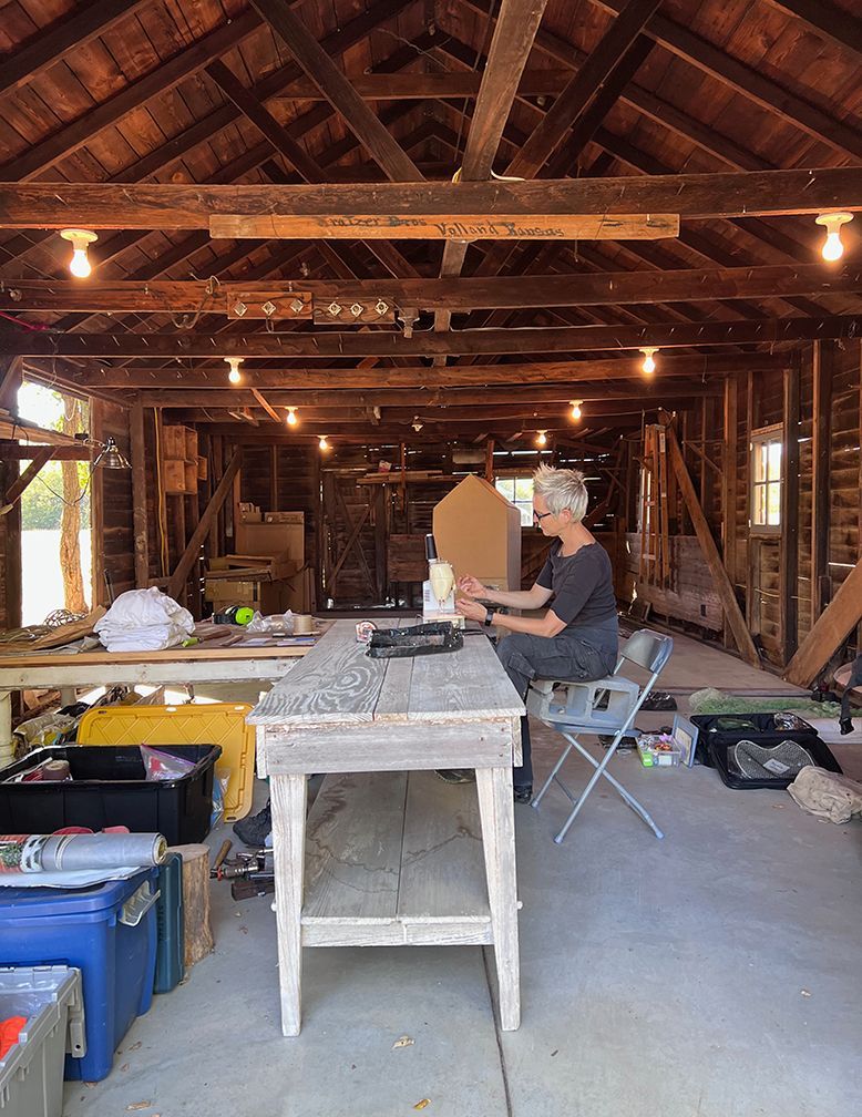 Person sitting at a weathered table inside a rustic, open-walled barn, working with papers.