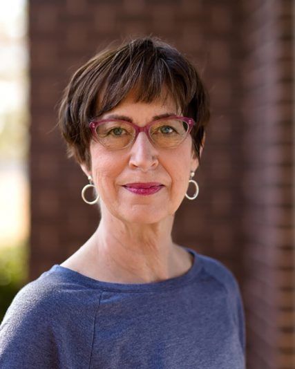 Woman with short brown hair, glasses, and earrings in a blue shirt, smiling. Brick wall background.