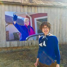 Woman in blue smiling, stands in front of a photo of herself waving.