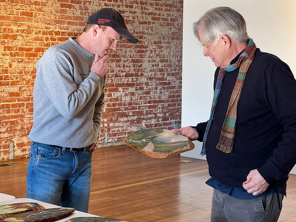 One man in a ball cap looking and considering an artwork that is held by the collector of log slab paintings.