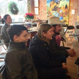 Group of people sitting and listening. A man wears a cowboy hat. Festive decorations are in the background.