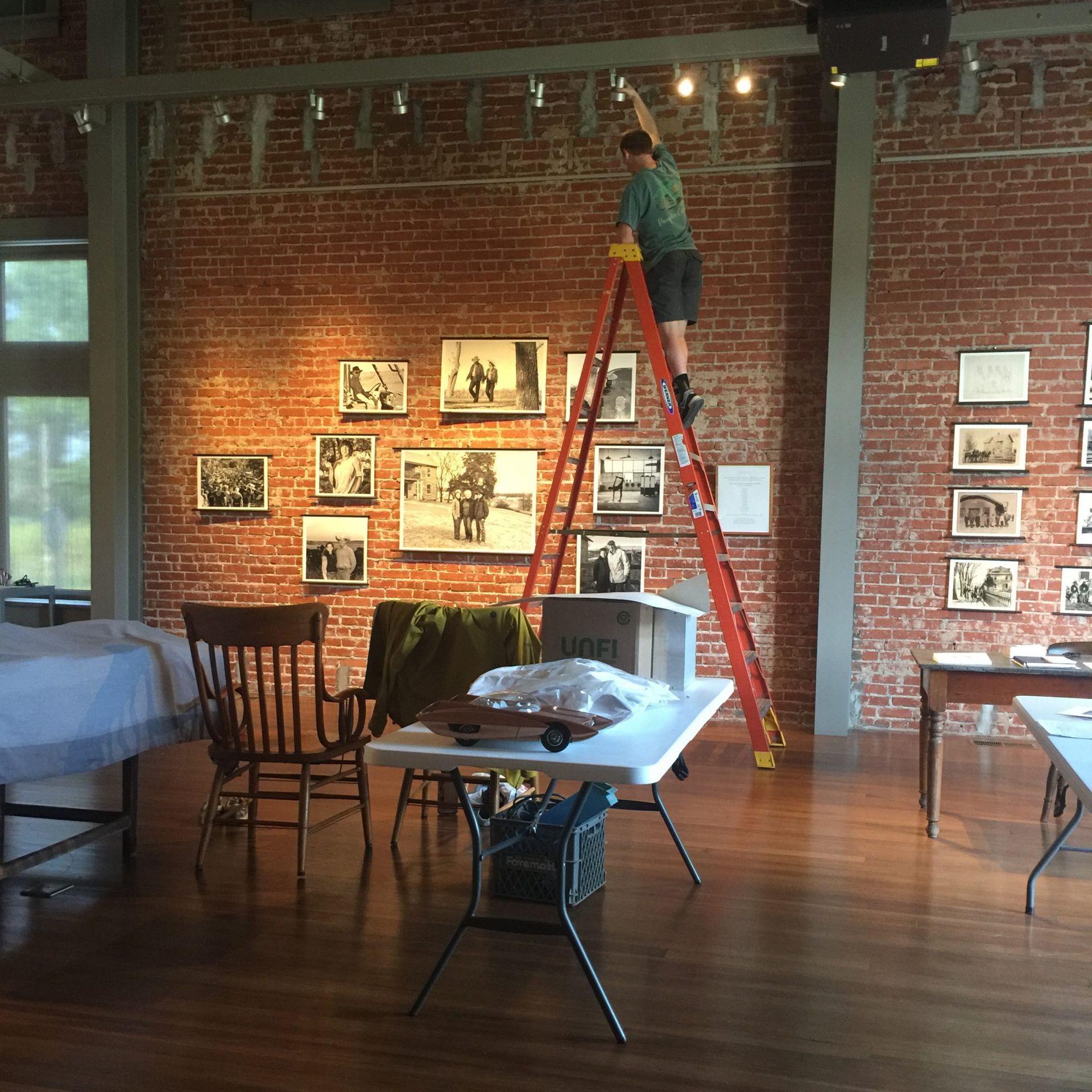 Man on a ladder adjusts a light in an art gallery. Brick wall, photos, wood floor, tables, and chairs are present.