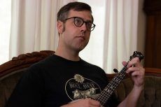 Man with glasses holding a banjo, sitting on a couch.