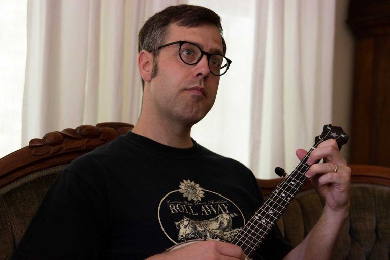 Man with glasses playing a banjo, sitting on a couch.