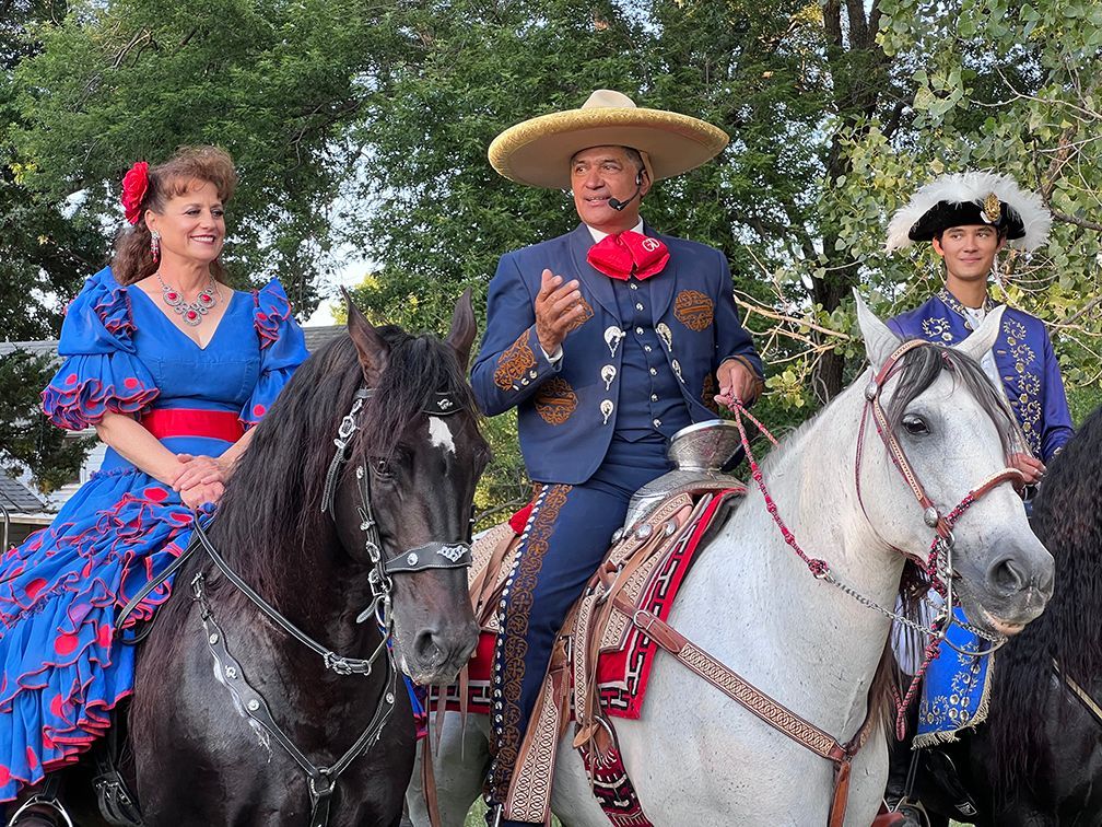 Three riders in traditional outfits on horses; man in sombrero speaks, woman in blue dress smiles.