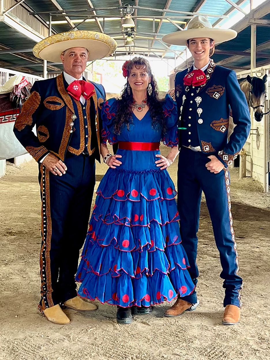 Three people in traditional Mexican attire pose in a stable. Two men wear charro suits, and a woman wears a blue dress.