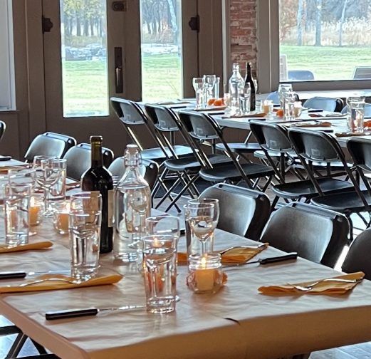 Long table set for a meal, glasses, candles, and water bottles, with outdoor view through windows.