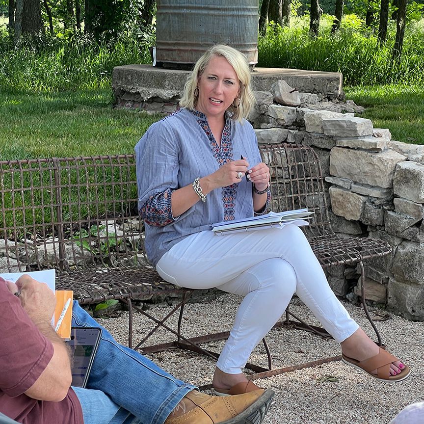 Woman in white pants and blue shirt sits outdoors, holding papers, talking.