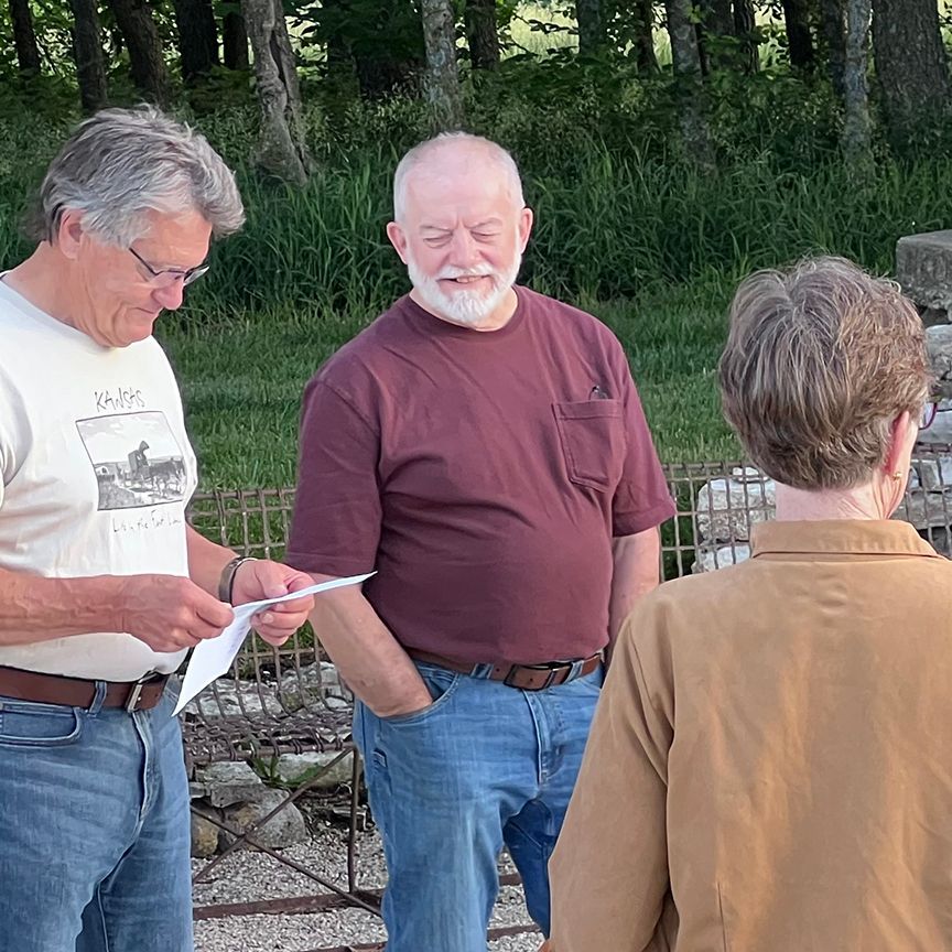 Three people outdoors. Man in white shirt reads paper to two others. Man with beard looks on. Woman in tan jacket is seen from behind.