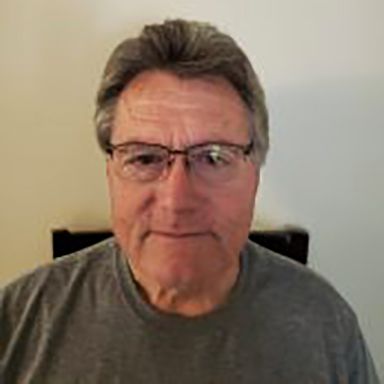 Man with gray hair and glasses wearing a gray shirt, indoors.