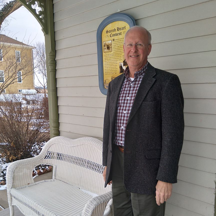 A man in a suit stands in front of a wicker bench