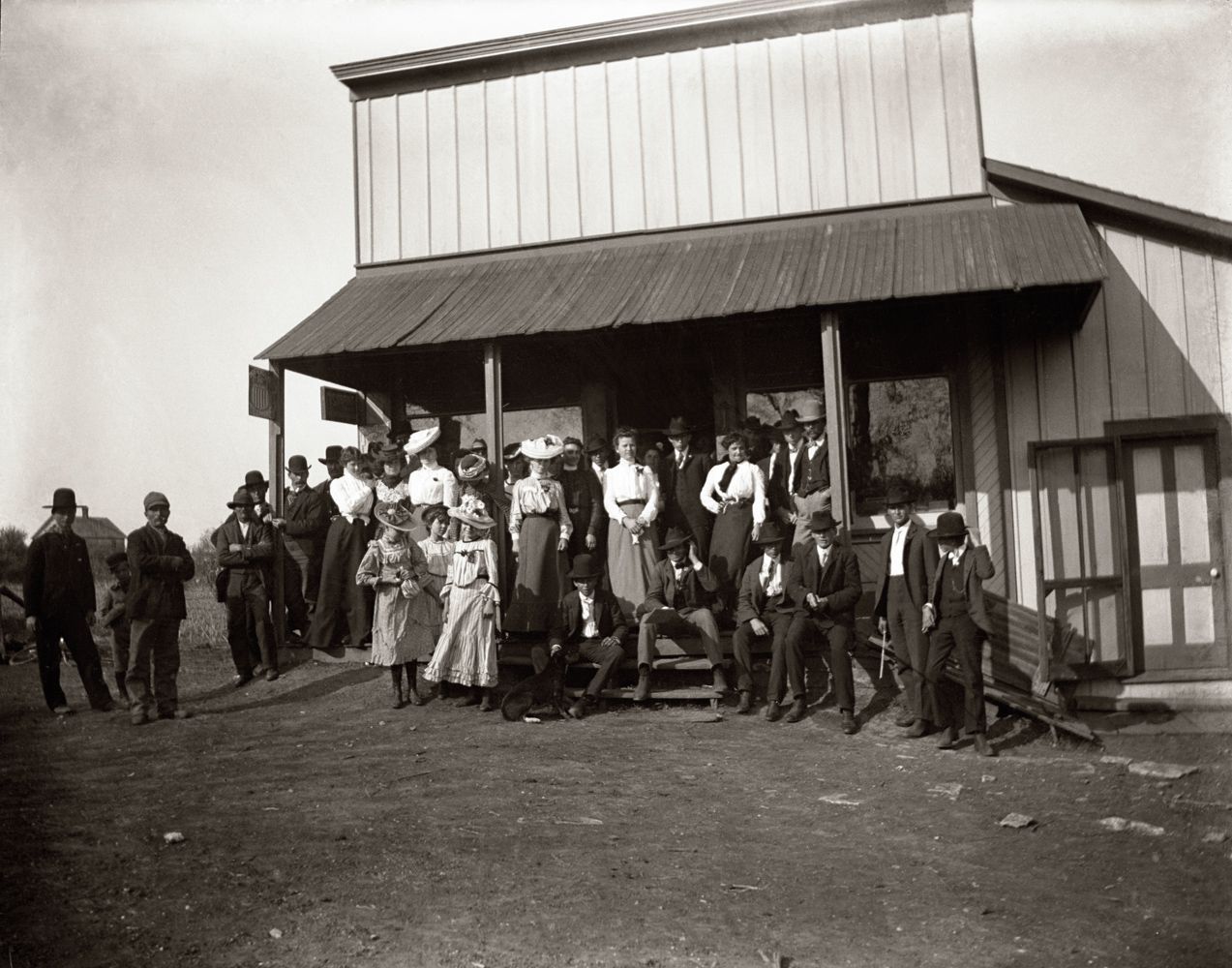 Group of people posed outside a wooden building with a porch, likely late 1800s. Men in suits and hats, women in long dresses.