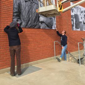 Two people installing a large black-and-white photo on a brick wall. One in a lift, one on ground level.