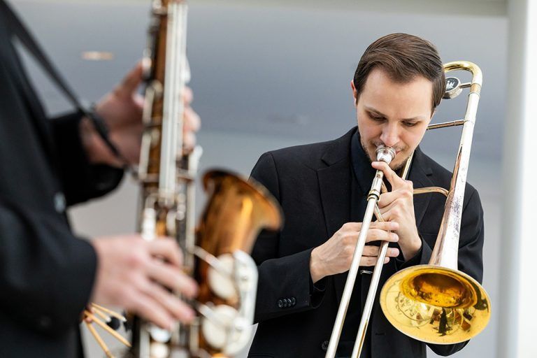 a person with light skin and brown hair plays the trombone outside. they wear a black suit