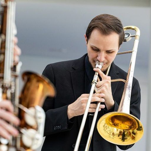 a person with light skin and brown hair plays the trombone outside. they wear a black suit