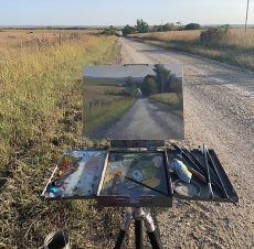 A painting of a dirt road is on an easel in a field.