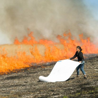 Woman unrolling a white sheet in front of a wildfire with orange flames and smoke.