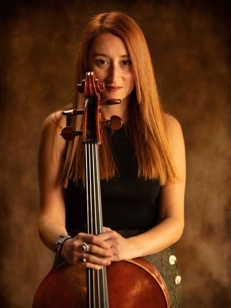 Woman with red hair holding a cello, looking at the viewer. Brown backdrop.