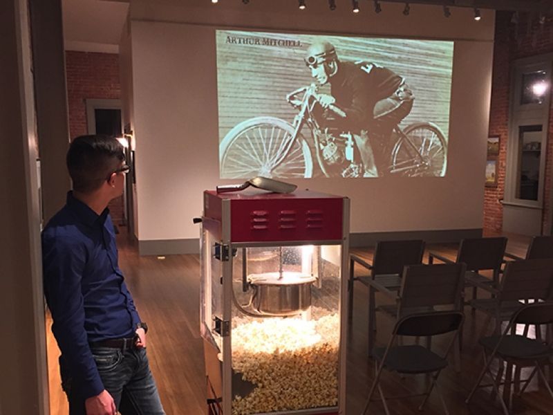 A man is standing in front of a popcorn machine with a picture of a man on a motorcycle projected on the wall.