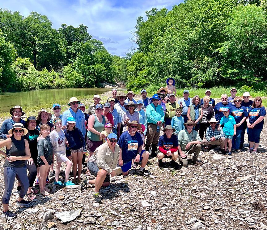 Large group of people standing along a riverbank with trees in the background on a sunny day.