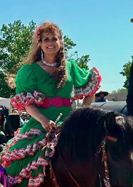 Woman in green dress with pink trim riding a horse in an outdoor parade, smiling.
