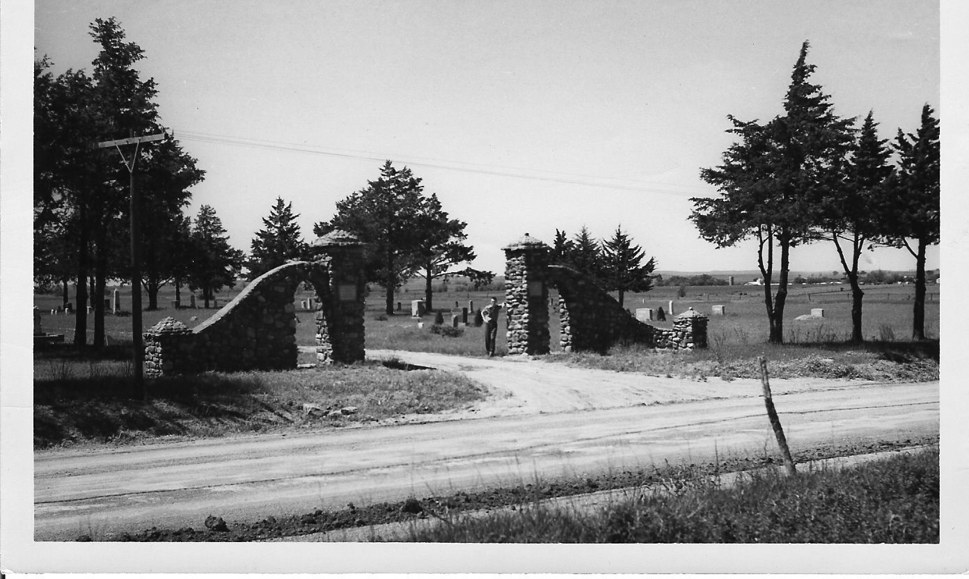 Stone entrance gates to a cemetery with a dirt road leading inside. Trees line the sides.