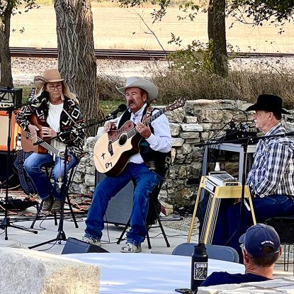 Three musicians playing guitars outdoors, wearing cowboy hats.