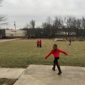 Girl in red jacket runs toward camera, other kids and train in background.