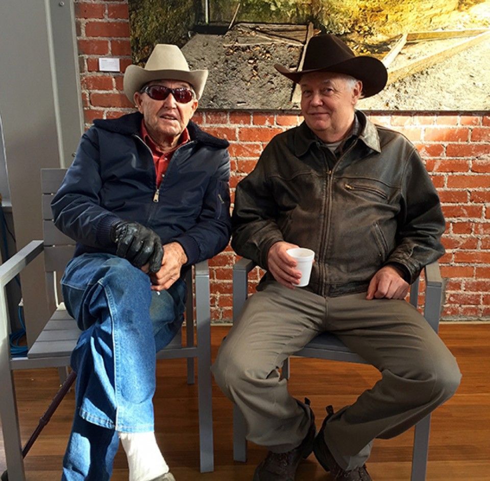 Two men in cowboy hats seated, smiling in front of artwork.