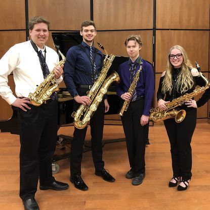 Four people with saxophones in a room with a piano. They are wearing formal attire and smiling.