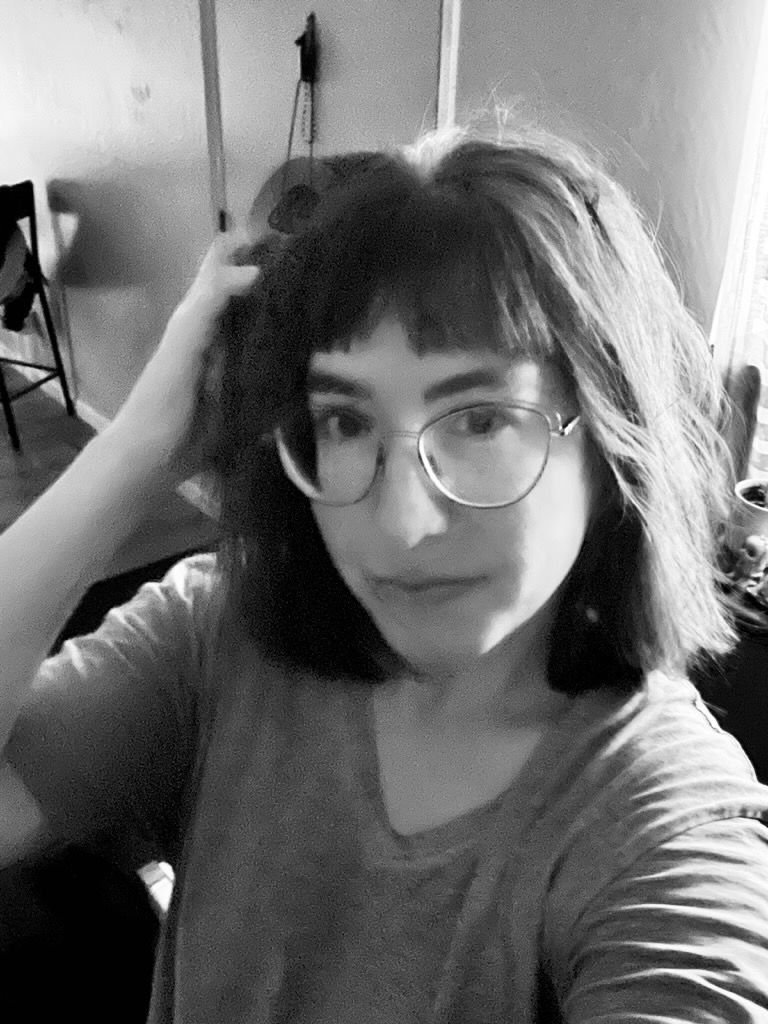 Woman with glasses, bangs, and gray shirt looking at the camera, hand in hair.